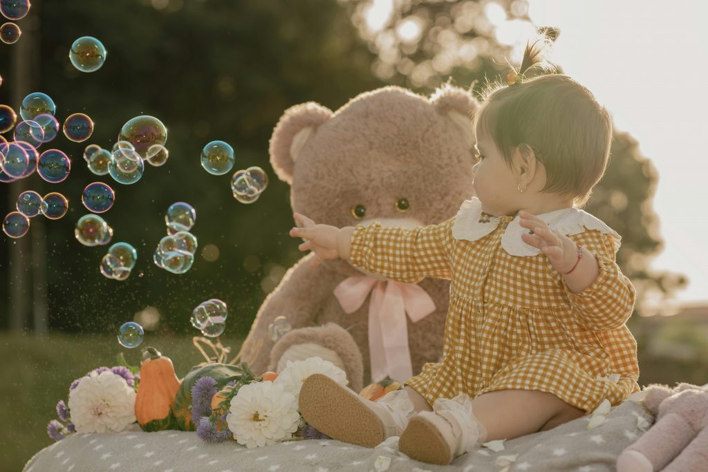 Charming outdoor scene with a baby interacting with bubbles and a teddy bear in soft sunlight.