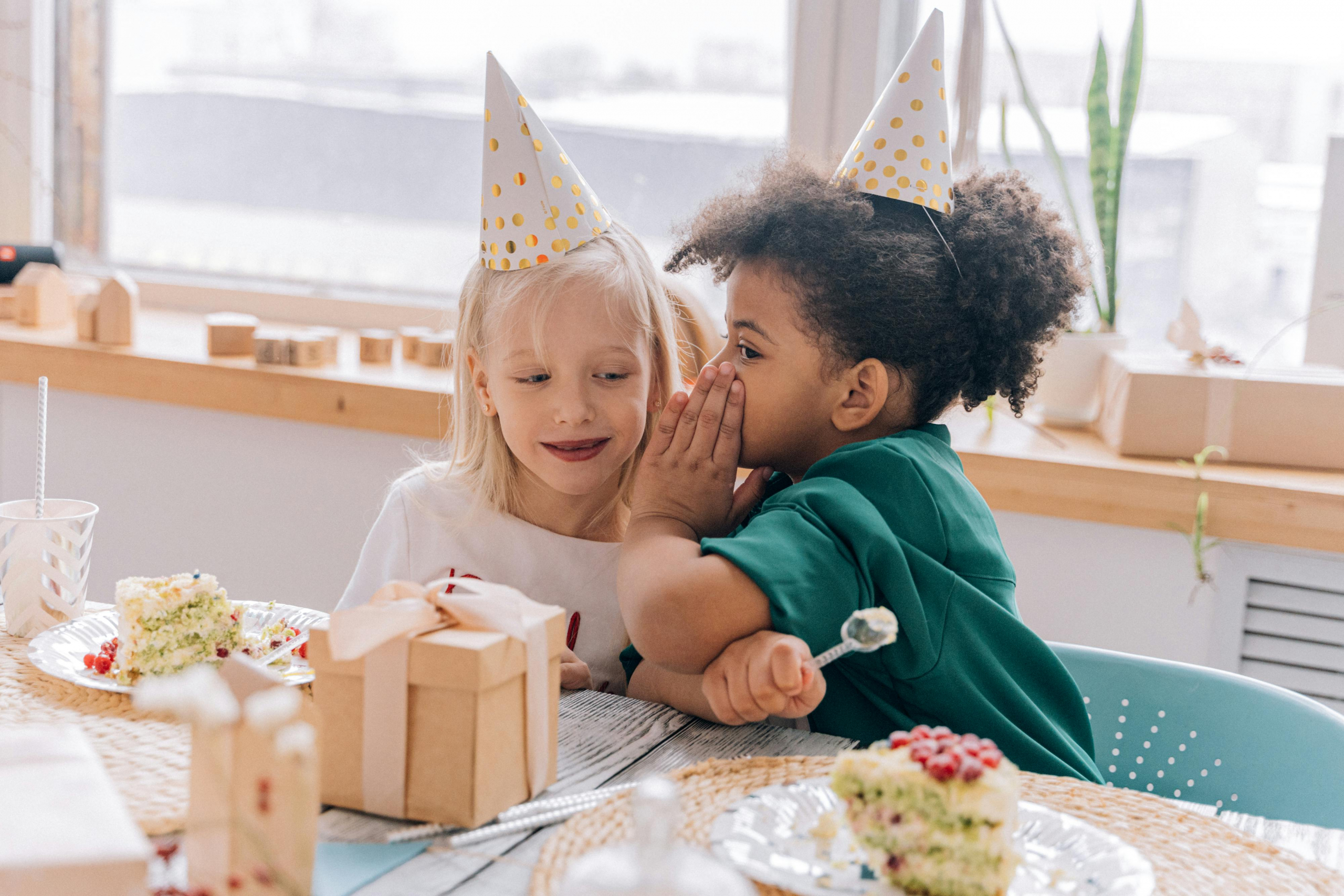 Two children wearing party hats, whispering and enjoying birthday cake indoors.