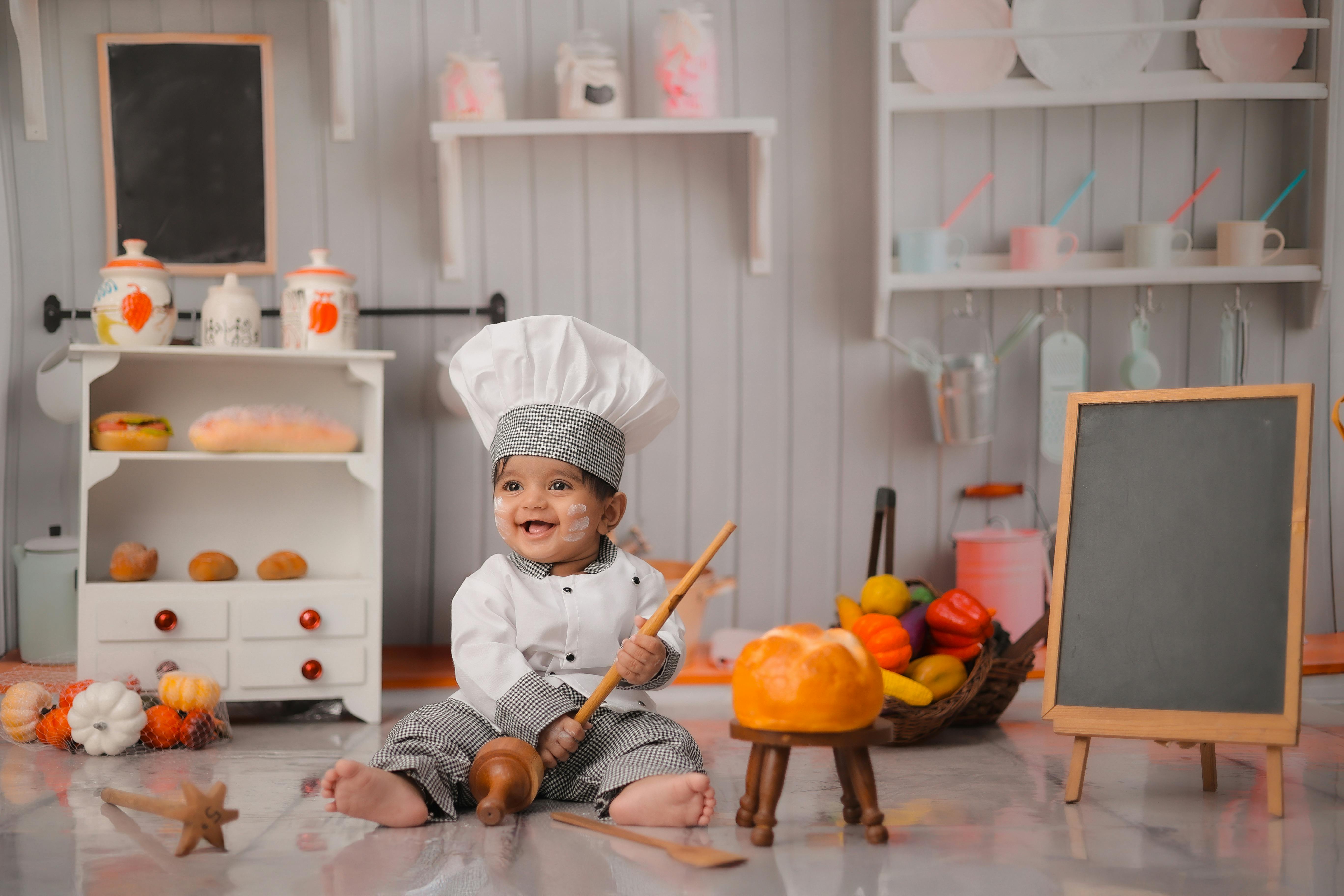 A cute baby dressed as a chef in a playful kitchen setting, surrounded by toy food and baking tools.