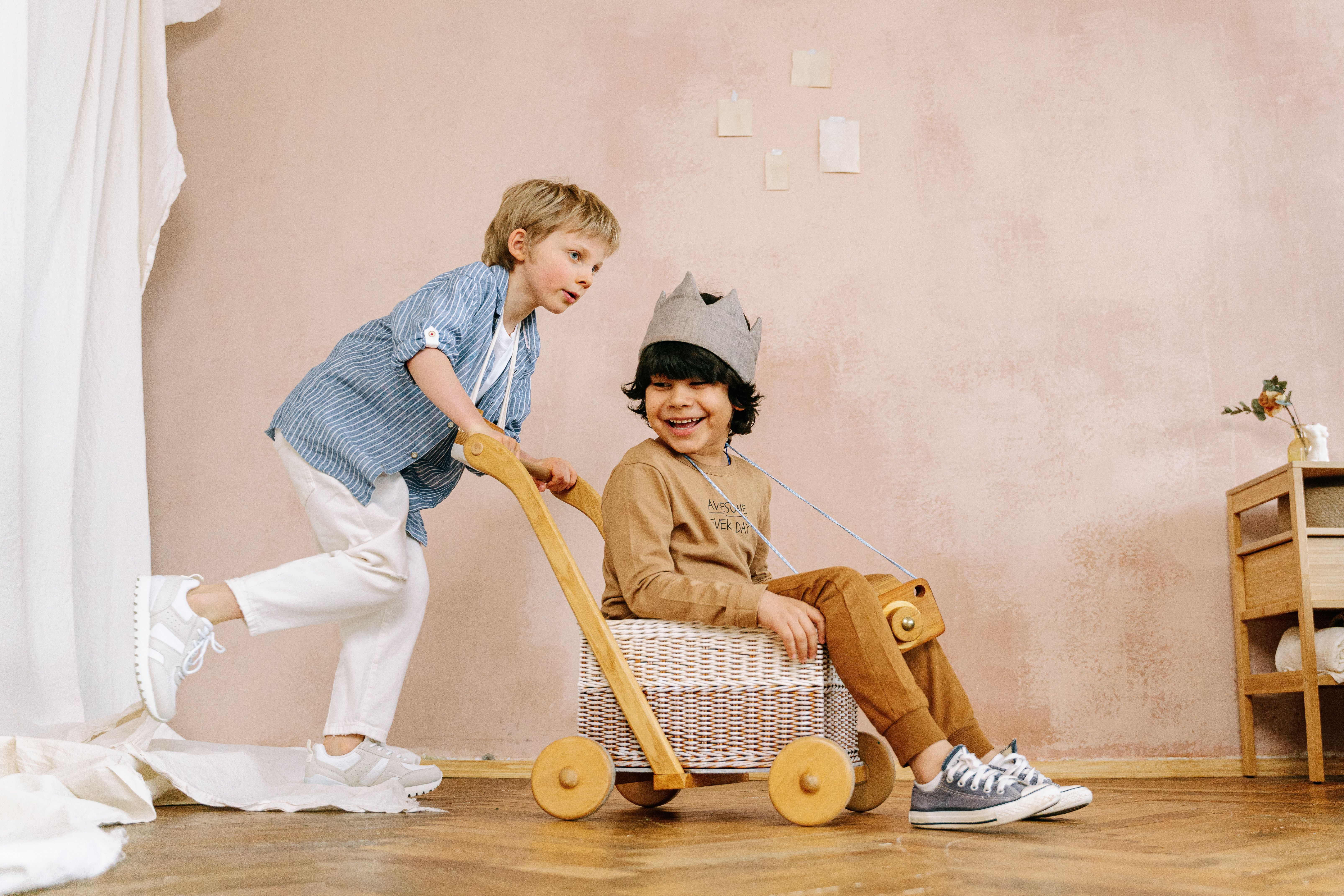 Two children playing with a handmade wooden toy cart indoors, smiling and enjoying themselves.