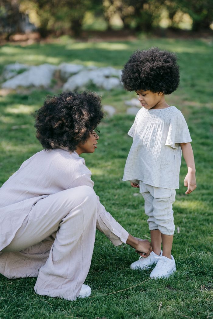 A mother ties her daughter's shoes on a grassy park field, showcasing a tender family moment.